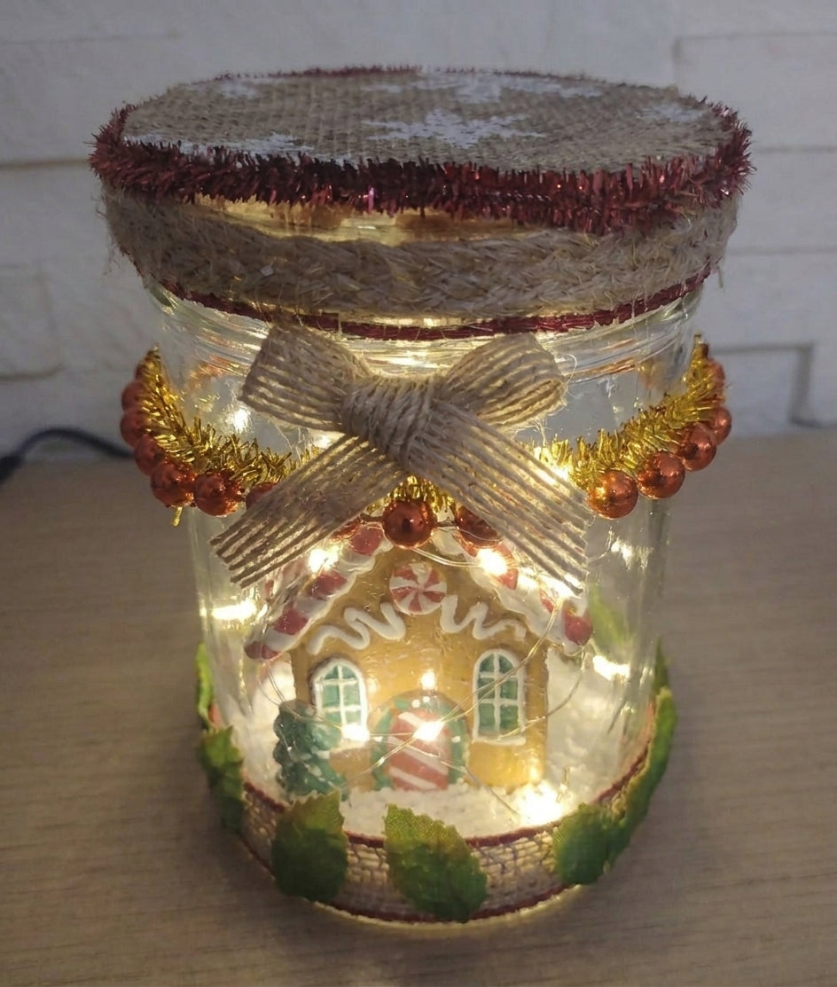 Lanterne de Noël en verre réutilisé illuminée, avec petite maison, sapins et couvercle décoré d’un nœud.